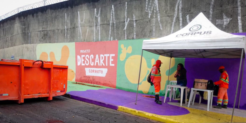 Em um espaço urbano com um grande muro ao fundo, há uma área colorida com grafismos e a inscrição "Projeto Descarte Correto". Uma caçamba laranja está posicionada na rua para coleta de materiais descartados. Ao lado, uma tenda branca com a marca "Corpus" abriga uma mesa e cadeiras, onde uma pessoa conversa com dois agentes de limpeza urbana, que vestem uniformes laranja com faixas refletivas. Outro agente está próximo à tenda. A cena retrata uma ação de conscientização e organização para o descarte correto de resíduos.